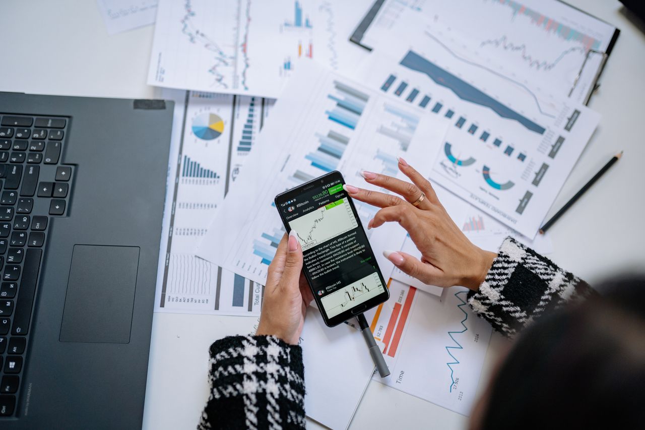 Businesswoman reviewing financial charts on a smartphone, with a laptop and documents nearby.